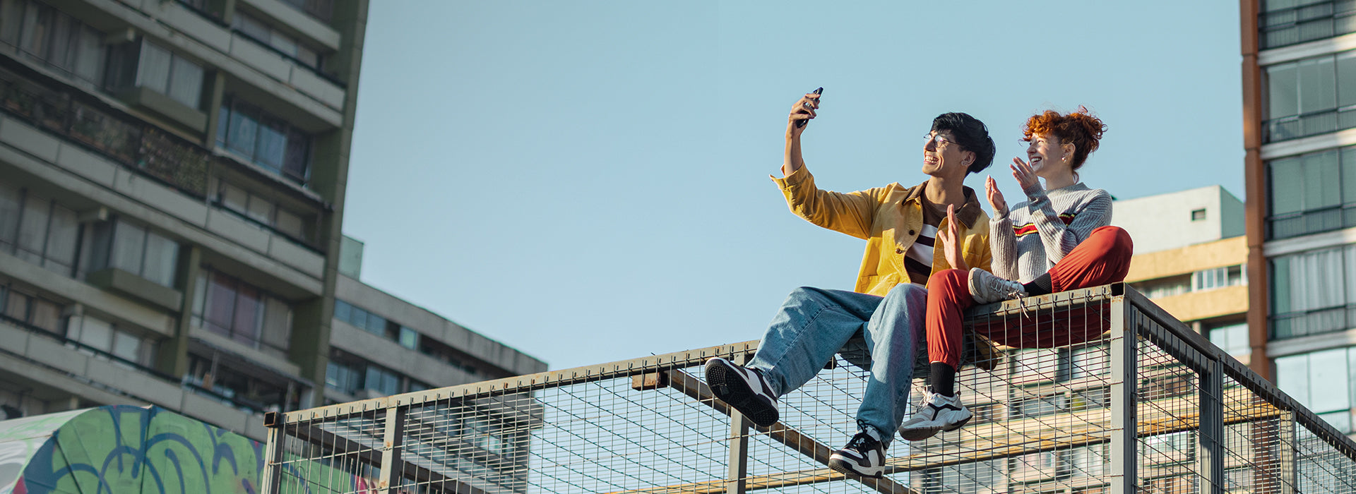 Two people sitting on a railing taking a selfie with a clear blue sky and urban buildings in the background.