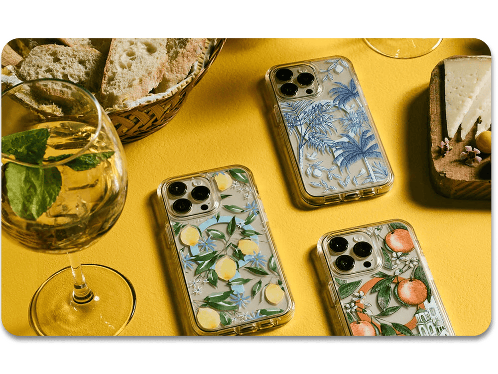 Three phone cases with floral and fruit designs on a yellow surface with bread and a glass.